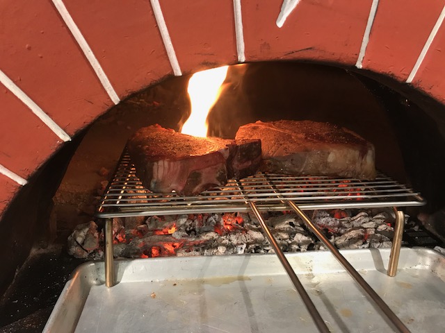 Our Keywords: A steak is being cooked in a wood-fired oven by our team.